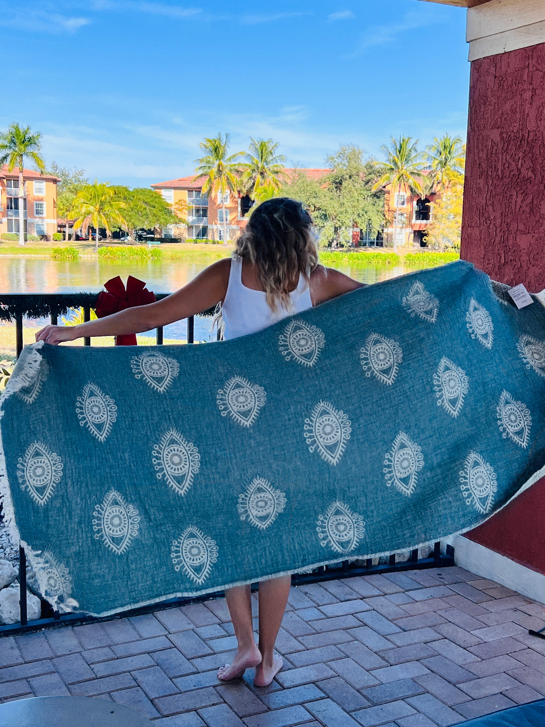 A woman stands on a patio holding the Eye Sage Sand Resistant Turkish Beach Towel by turkanhome.com, featuring white patterns. She faces a pond and palm trees with apartment buildings in the background under a bright blue sky.
