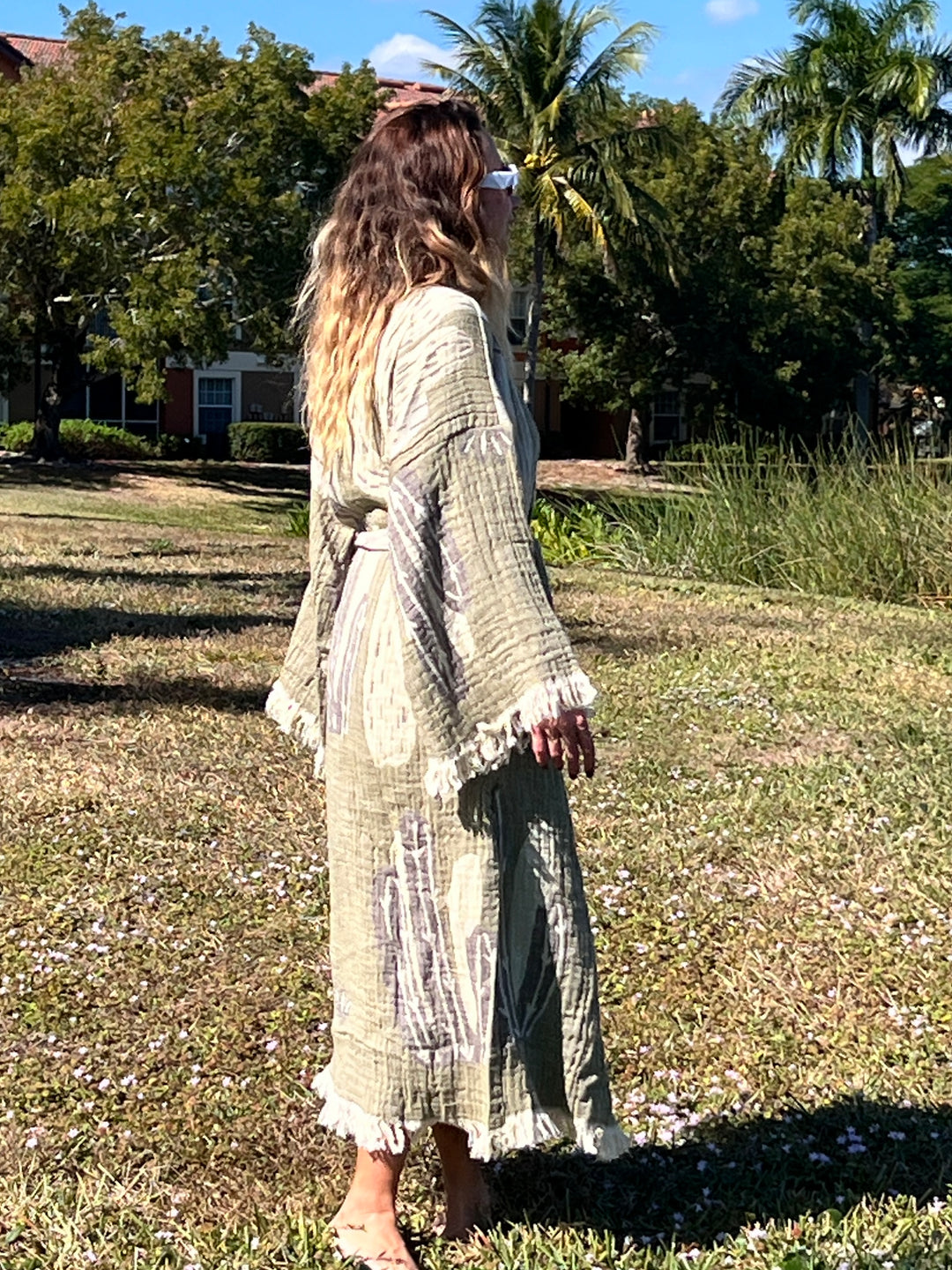 A woman with long wavy hair and sunglasses models the light green Cactus Robe by turkanhome.com as lounge or beach wear, standing barefoot on grass in a sunny outdoor setting with trees and buildings in the background.