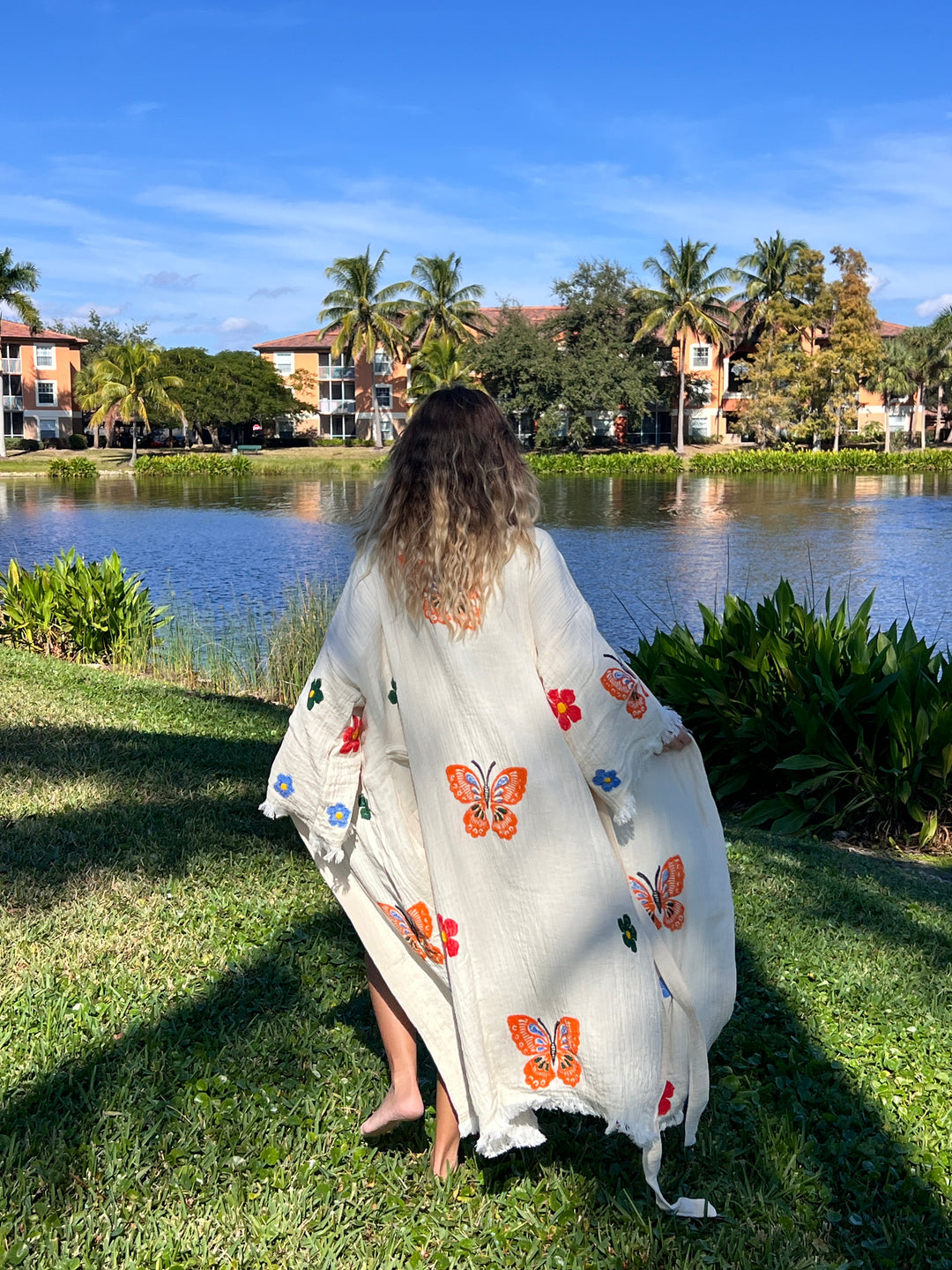 A person with long wavy hair wears the Butterfly Kimono Robe-Natural Color from turkanhome.com, decorated with colorful butterflies and flowers, standing by a lakeside with palm trees and apartments under a bright blue sky.