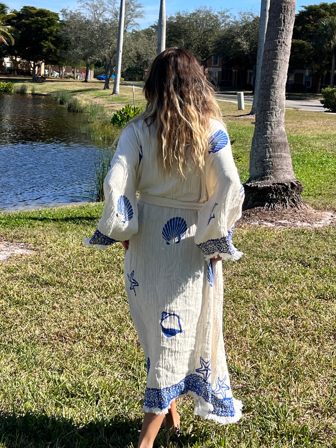 A woman with long, wavy hair stands barefoot on grass by a pond, wearing the Sea Shell Kimono Robe-Blue from turkanhome.com. She faces away from the camera; trees and houses are visible in the background.