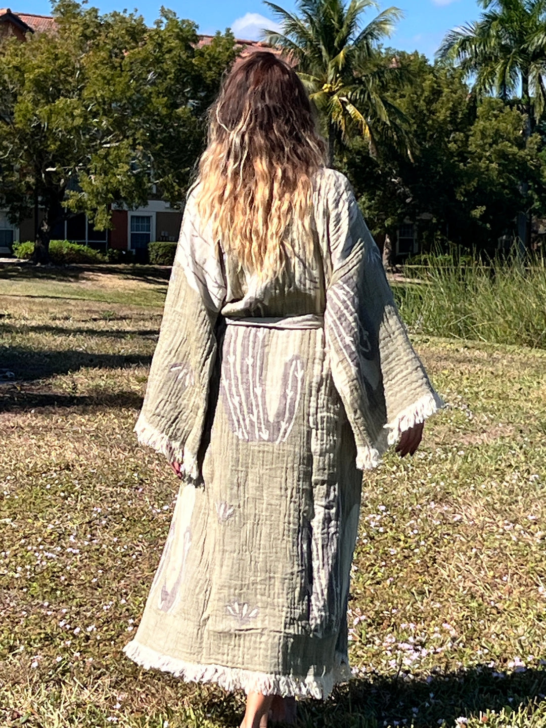 A person with long, wavy hair stands barefoot on grass outdoors, facing away, wearing the Cactus Robe from turkanhome.com—a light green cotton muslin kimono—amid palm trees and a building under a sunny sky.