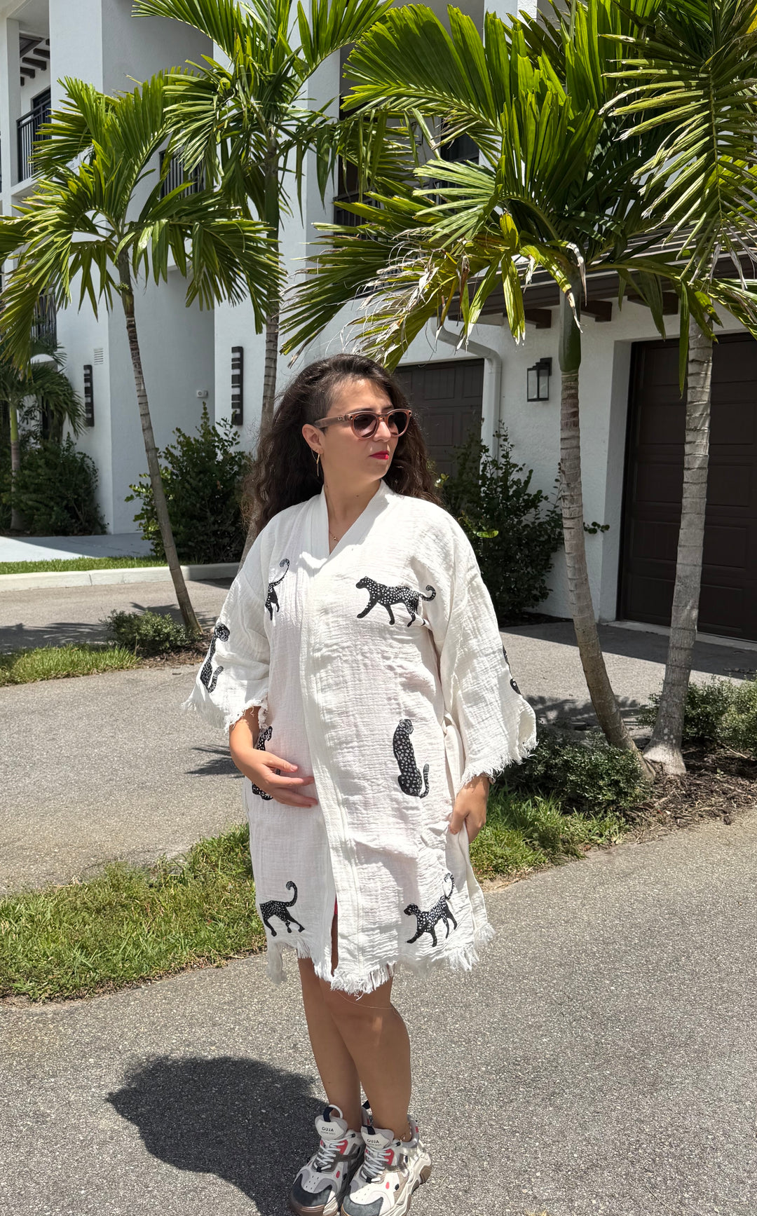 A woman with long dark hair in sunglasses wears a white dress with black animal prints and sneakers, standing outside near palm trees and a white building on a sunny day, her effortlessly chic look completed by the Leopard Cotton Kimono Robe-Short from turkanhome.com draped over her shoulders.