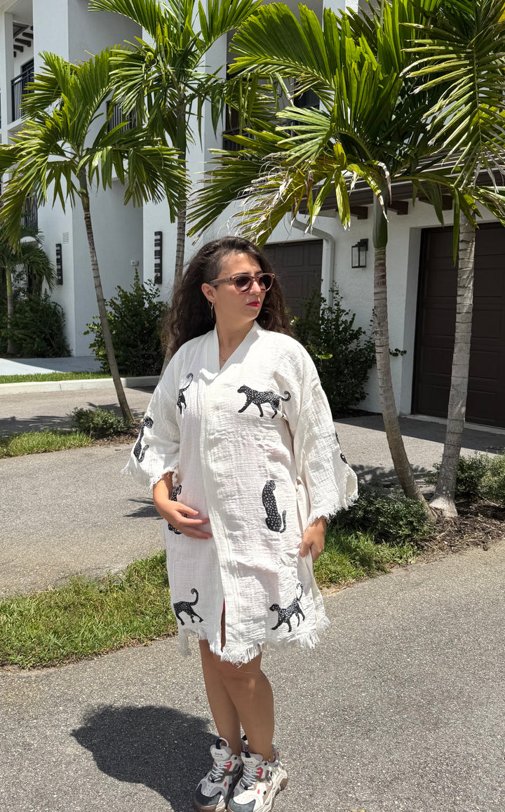 A woman with long dark hair in sunglasses wears a white dress with black animal prints and sneakers, standing outside near palm trees and a white building on a sunny day, her effortlessly chic look completed by the Leopard Cotton Kimono Robe-Short from turkanhome.com draped over her shoulders.