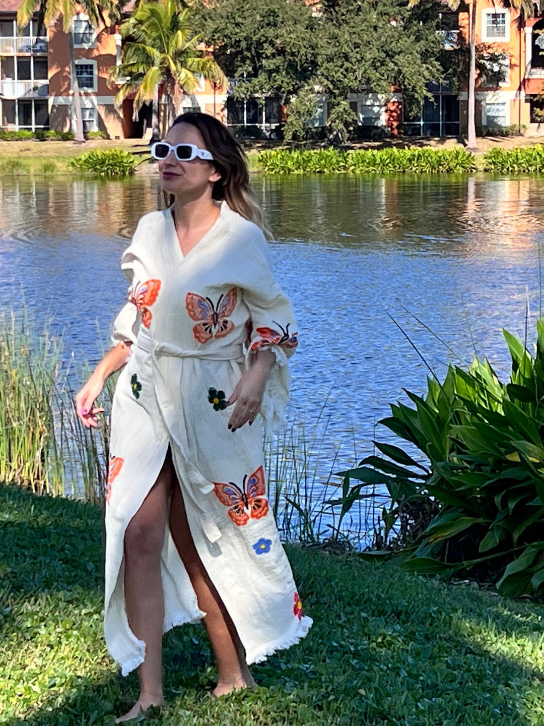 A woman stands barefoot on grass by a lake wearing the Butterfly Kimono Robe-Natural Color from turkanhome.com, featuring colorful butterfly and flower designs. She pairs it with white sunglasses; trees and apartments are visible in the background.