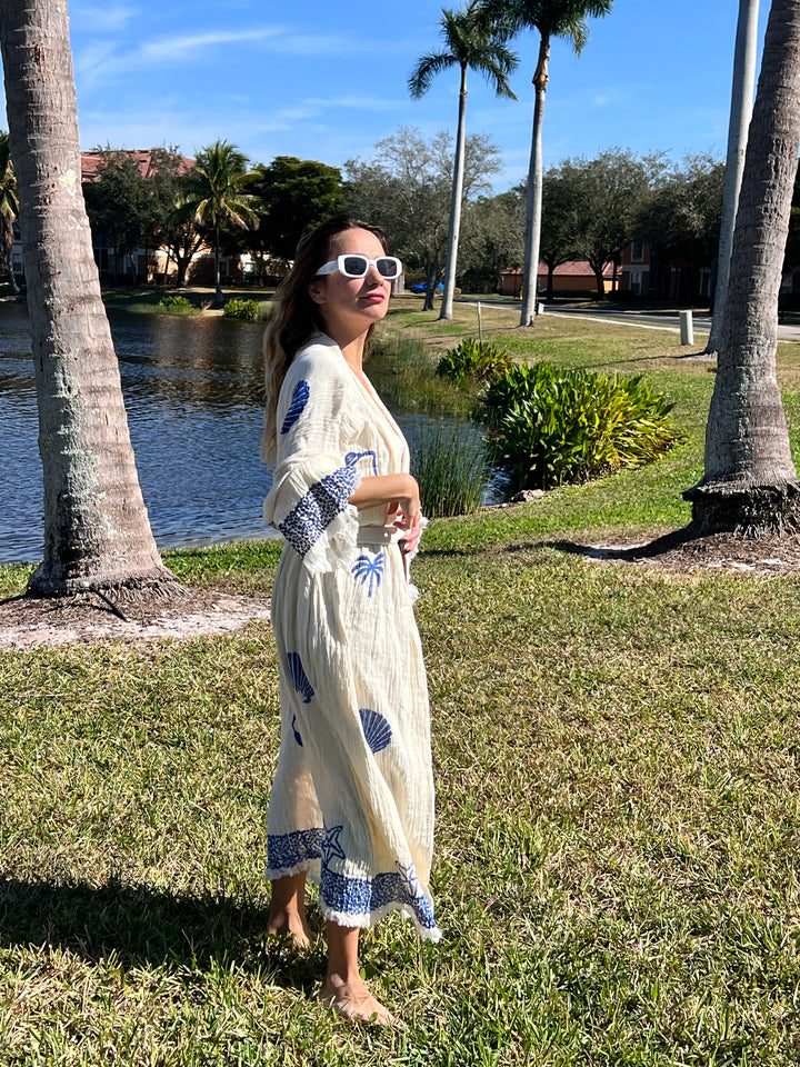 A woman in the Sea Shell Kimono Robe-Blue by turkanhome.com, sunglasses on, walks barefoot on grass near a lake with palm trees under a clear blue sky.