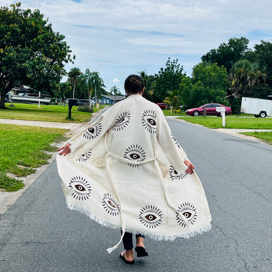 A person walks down a suburban street in the Brown Eye Kimono Robe from turkanhome.com, featuring large black eye designs. Their back faces the camera as they stroll past green lawns and leafy trees.
