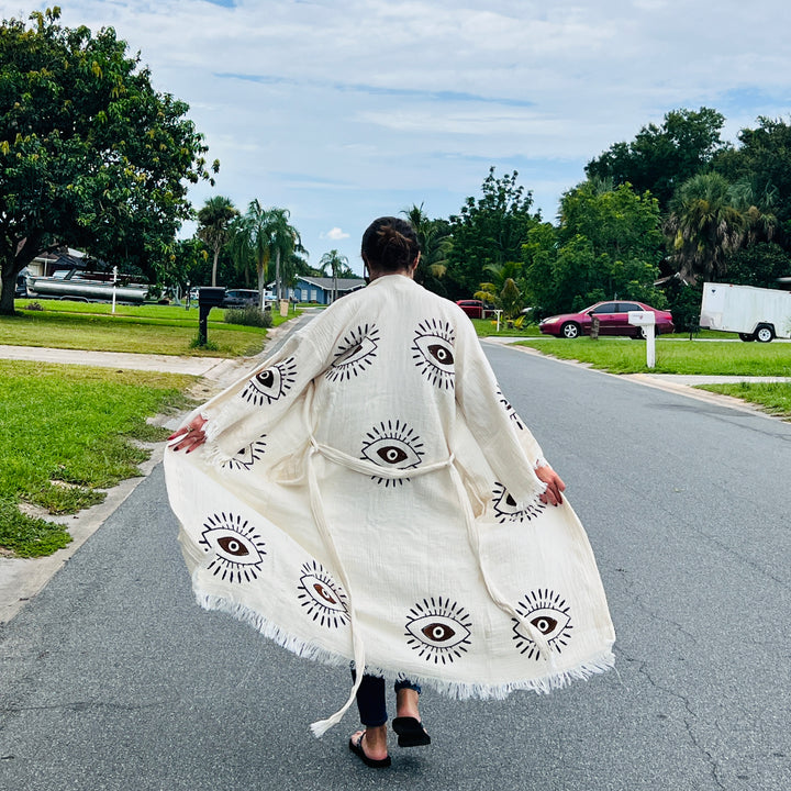 A person walks down a suburban street in the Brown Eye Kimono Robe from turkanhome.com, featuring large black eye designs. Their back faces the camera as they stroll past green lawns and leafy trees.
