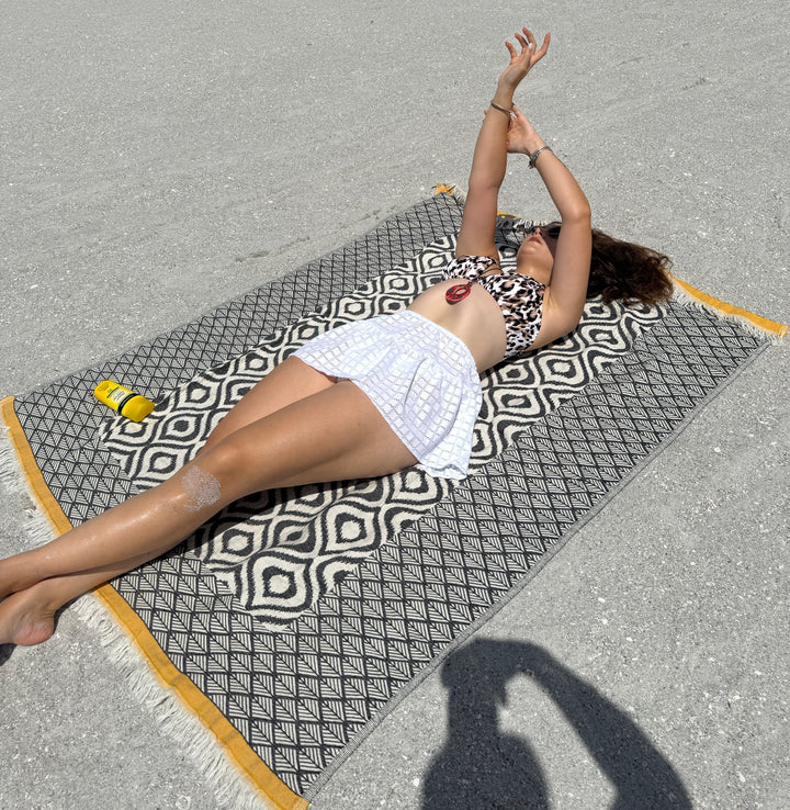 A woman in a patterned bikini top and white skirt lies on a turkanhome.com Ikat Throw with a reversible geometric pattern on the sand, arms stretched above her head. A yellow sunscreen bottle is beside her and the photographer's shadow is visible.