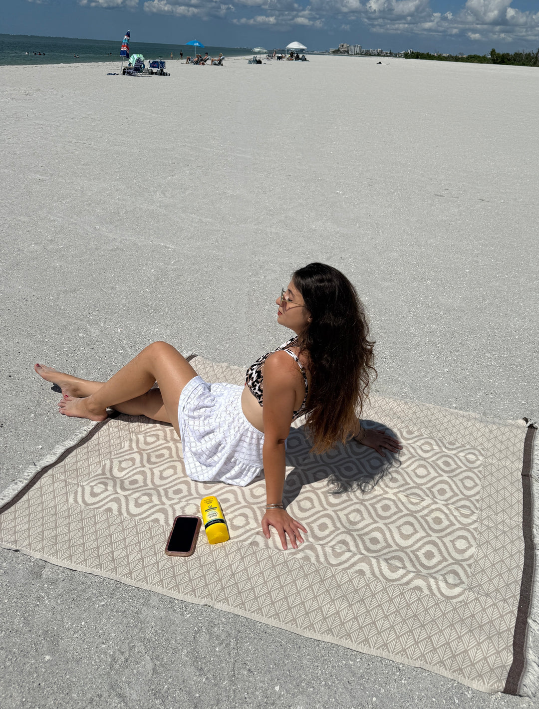 A woman in a white skirt and patterned bikini top relaxes on a sandy, mostly empty beach atop the Ikat Throw from turkanhome.com, featuring a reversible geometric design. Nearby are her phone and two yellow sunscreen bottles under the blue sky.