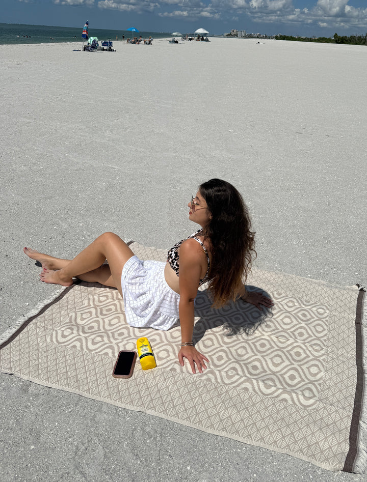A woman in a white skirt and patterned bikini top relaxes on a sandy, mostly empty beach atop the Ikat Throw from turkanhome.com, featuring a reversible geometric design. Nearby are her phone and two yellow sunscreen bottles under the blue sky.