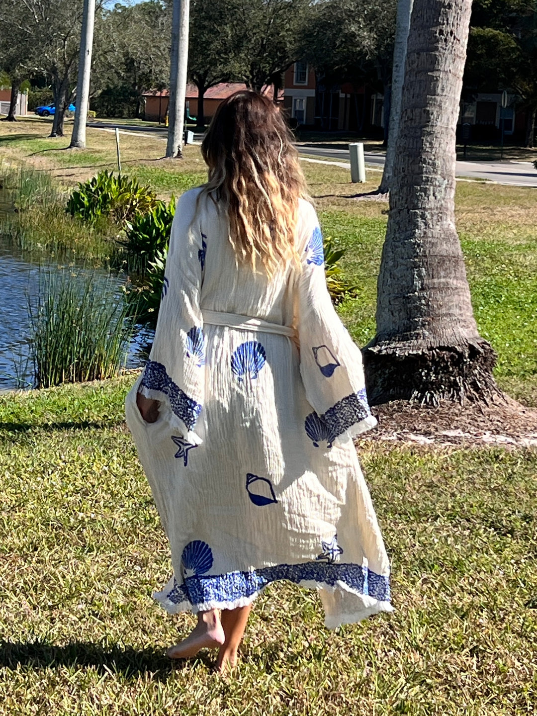 A woman with long wavy hair wears the Sea Shell Kimono Robe-Blue by turkanhome.com, walking barefoot on grass near a pond and palm tree, with houses and trees in the background.