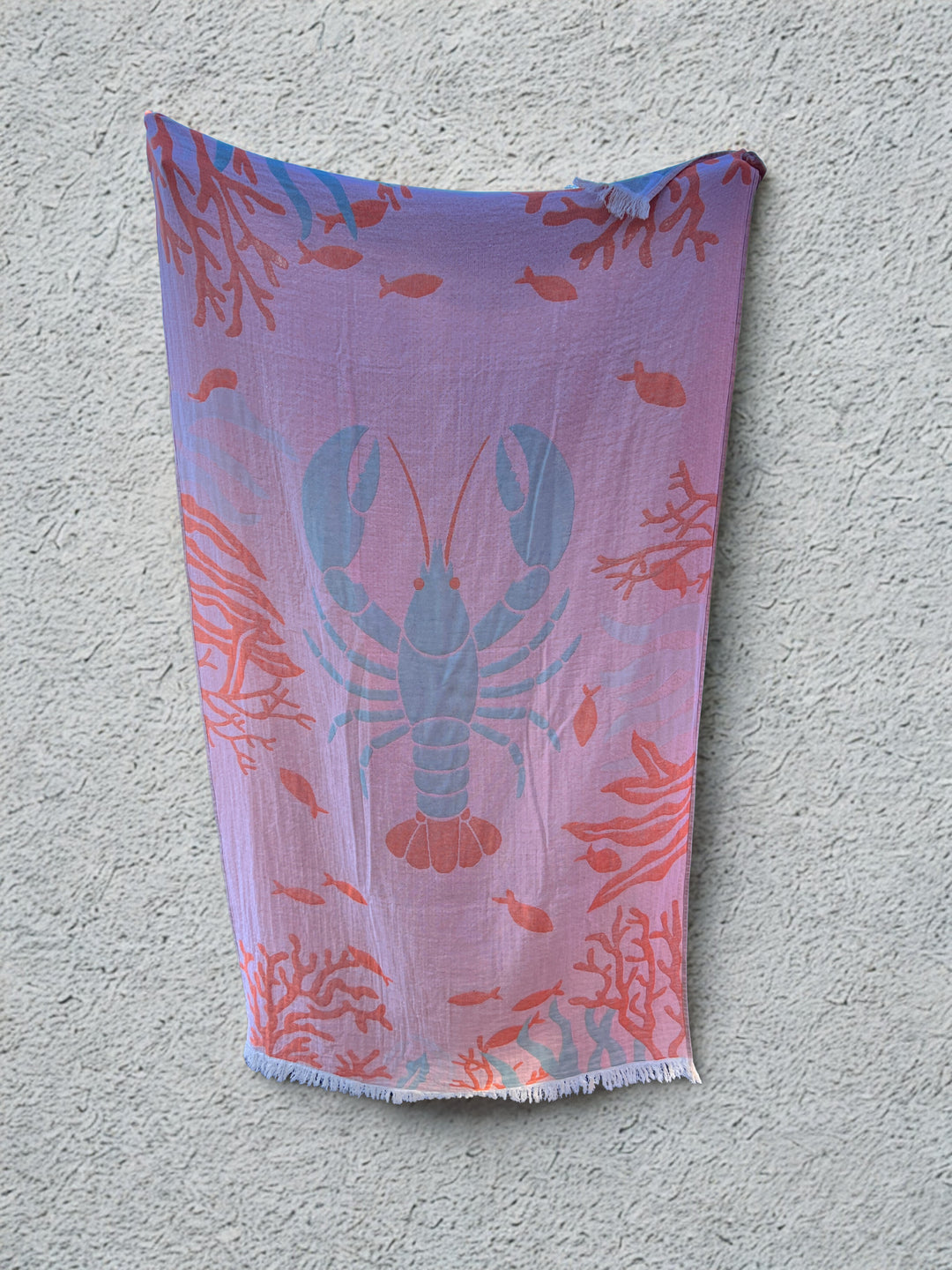 The Lobster Turkish Towel – Quick Drying Beach Towel by turkanhome.com features a reversible blue lobster design with red coral and small fish, hanging against a light gray wall. Made from soft Turkish cotton for ultimate comfort.