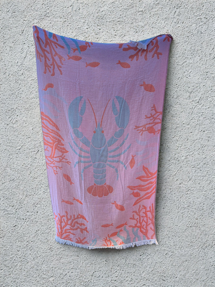 The Lobster Turkish Towel – Quick Drying Beach Towel by turkanhome.com features a reversible blue lobster design with red coral and small fish, hanging against a light gray wall. Made from soft Turkish cotton for ultimate comfort.