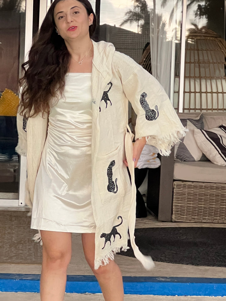 A woman with long dark hair wears a white satin dress and the Black Leopard Hooded Cotton Kimono Robe by turkanhome.com, featuring black panther designs. She stands on a patio with wicker furniture, glass doors, and palm trees reflected behind her.