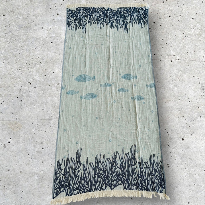 The Fishes and Coral Turkish Towel by turkanhome.com is a rectangular, light greenish-blue Jacquard-woven 100% cotton towel featuring dark blue coral at both ends and swimming fish in the center, displayed on speckled concrete.