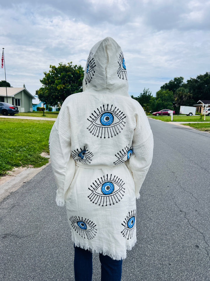 A person wearing the Blue Eye With Lashes Kimono Robe from turkanhome.com stands on a suburban street, facing away. Houses, green grass, and a cloudy sky create a peaceful backdrop for this stylish short loungewear look.
