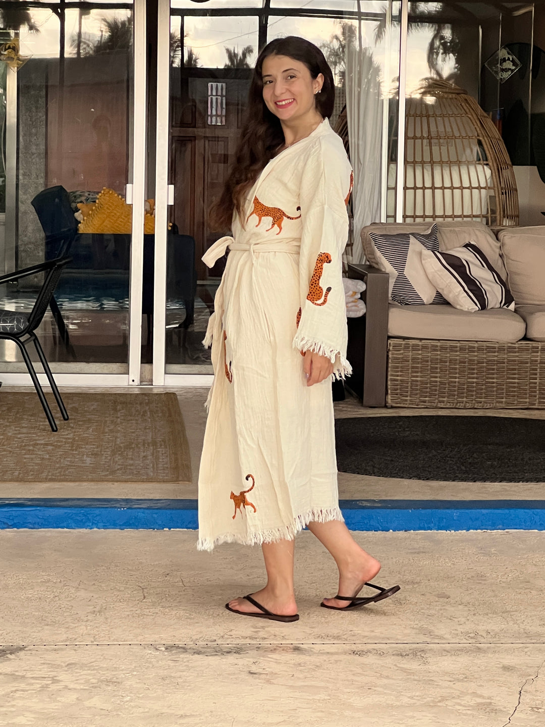 A woman with long hair smiles outdoors on a patio near a sofa and glass doors, wearing the Orange Leopard Cotton Kimono Robe – Lightweight Boho Beach Cover Up by turkanhome.com and black flip-flops.