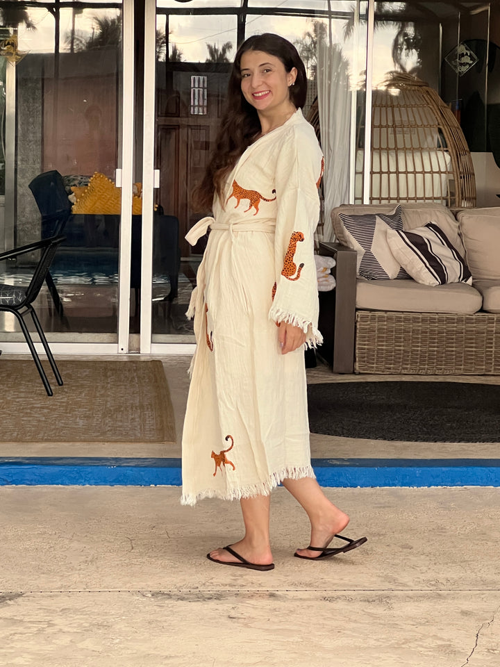 A woman with long hair smiles outdoors on a patio near a sofa and glass doors, wearing the Orange Leopard Cotton Kimono Robe – Lightweight Boho Beach Cover Up by turkanhome.com and black flip-flops.