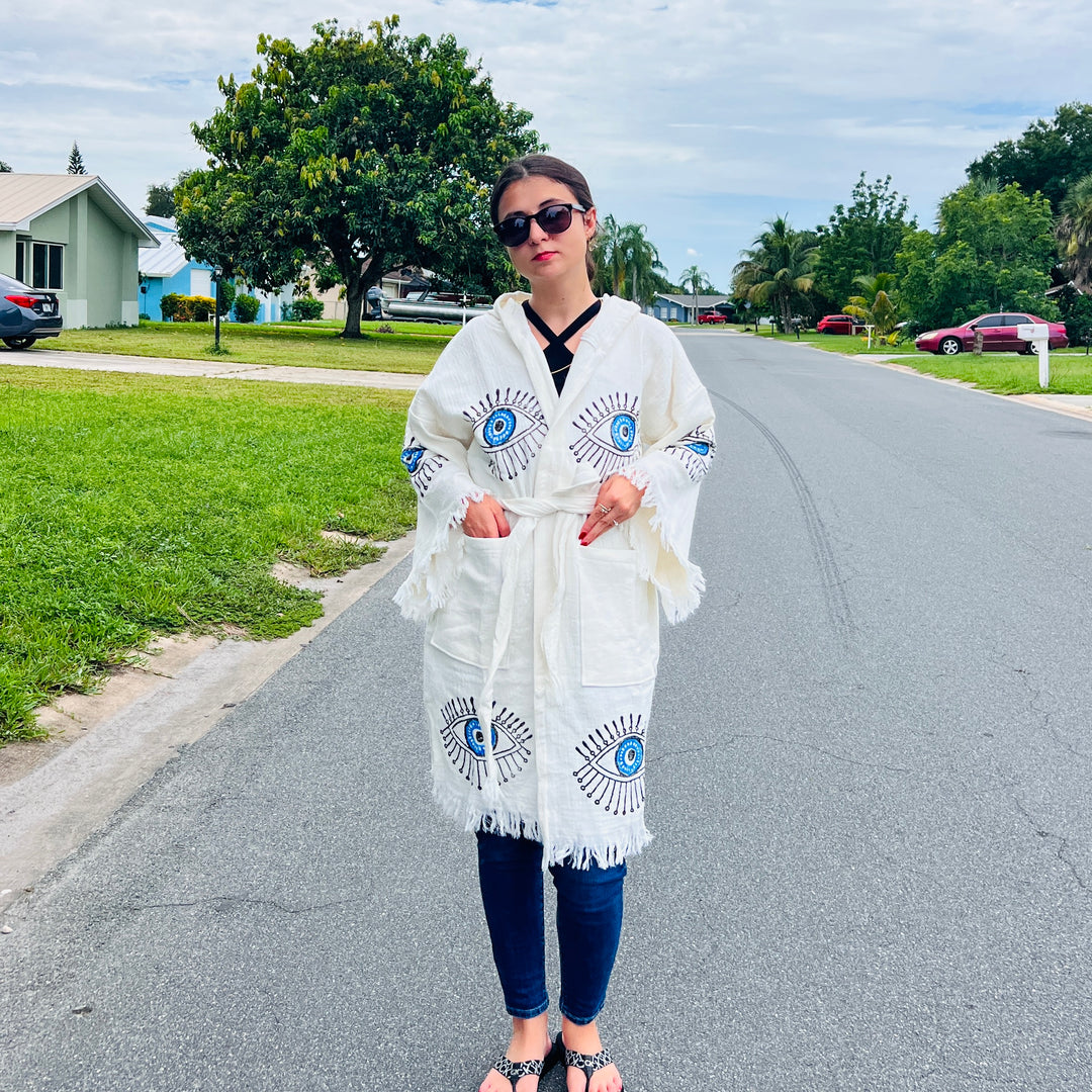 A woman stands on a quiet suburban street wearing sunglasses, jeans, sandals, and the Blue Eye With Lashes Kimono Robe (short) from turkanhome.com. Houses, trees, and grass line the peaceful neighborhood.