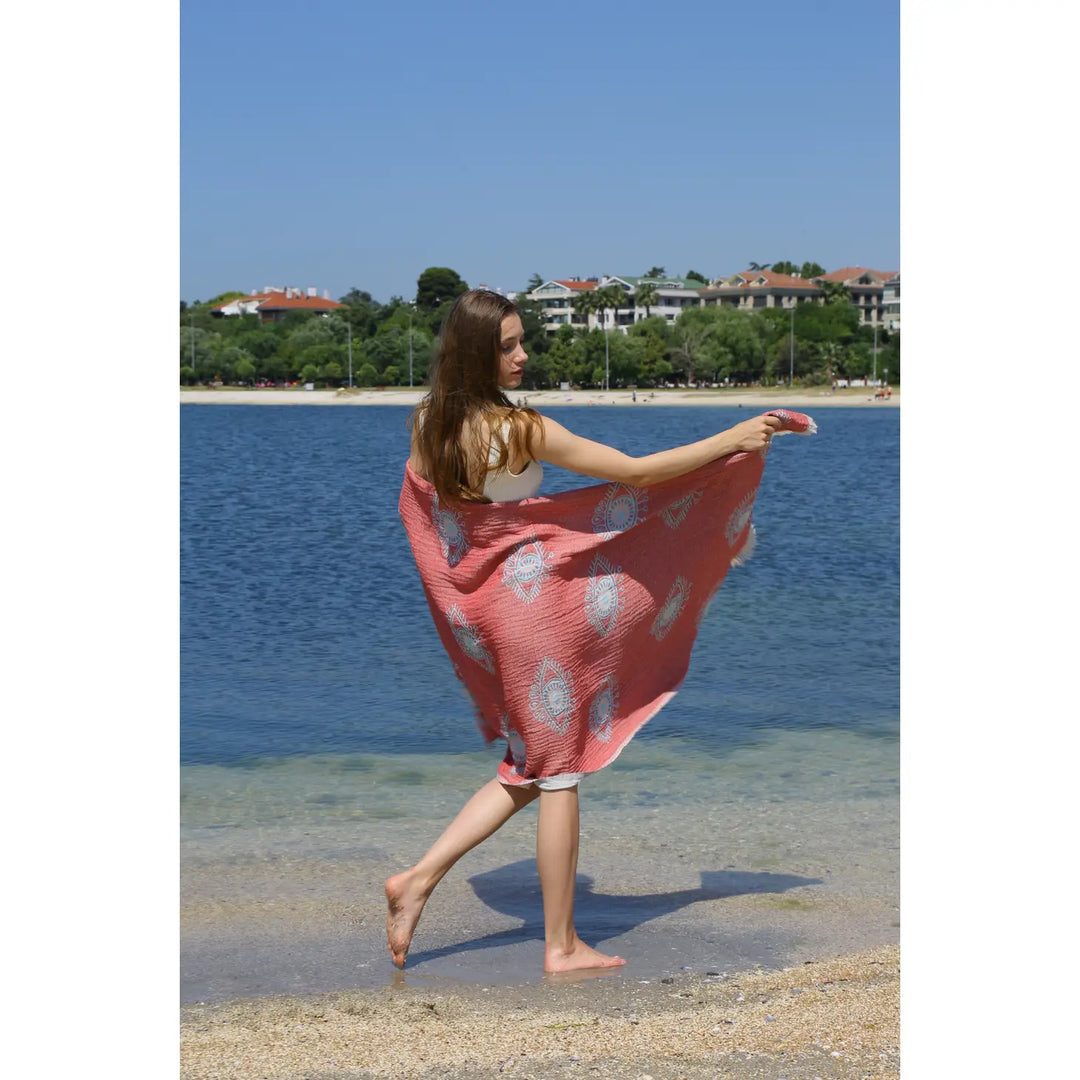 A woman stands barefoot on sand, holding the Eye Red Sand Resistant Turkish Beach Towel by turkanhome.com behind her. She faces calm water and distant houses under a clear blue sky.