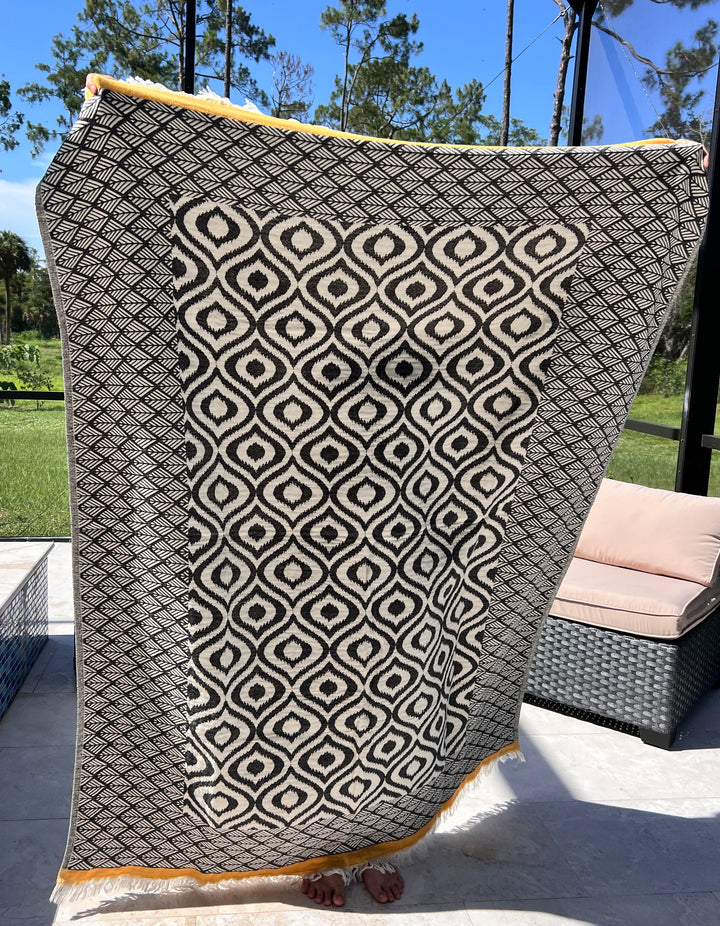 A person stands on a patio holding up the Ikat Throw from turkanhome.com, its large black-and-white geometric pattern hiding their body except for their toes. Outdoor furniture and green trees are in the background.