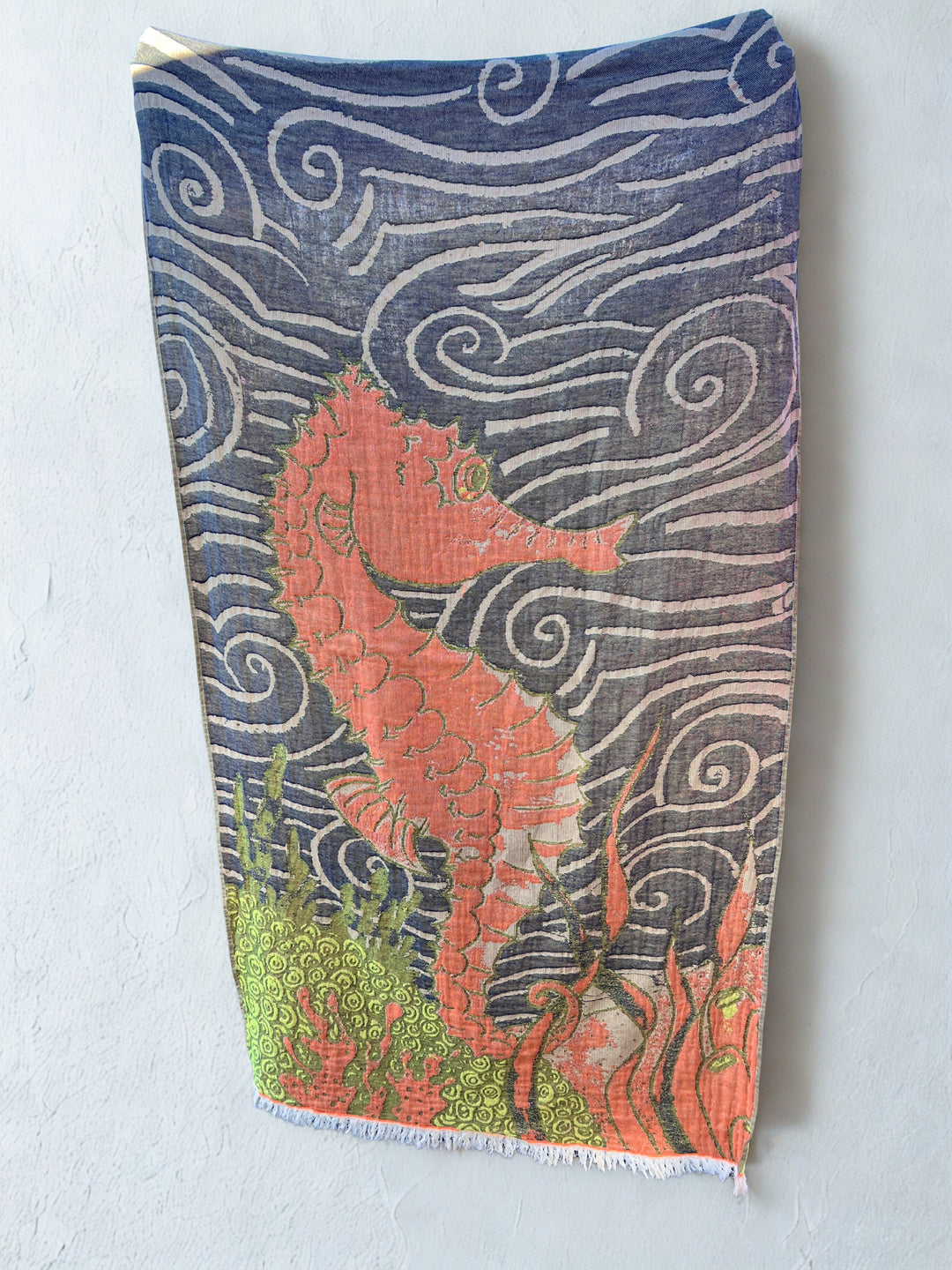 The Seahorse Turkish Towel from turkanhome.com, made of 100% cotton, features a bold red seahorse among blue swirls and green seaweed and hangs against a light gray wall.