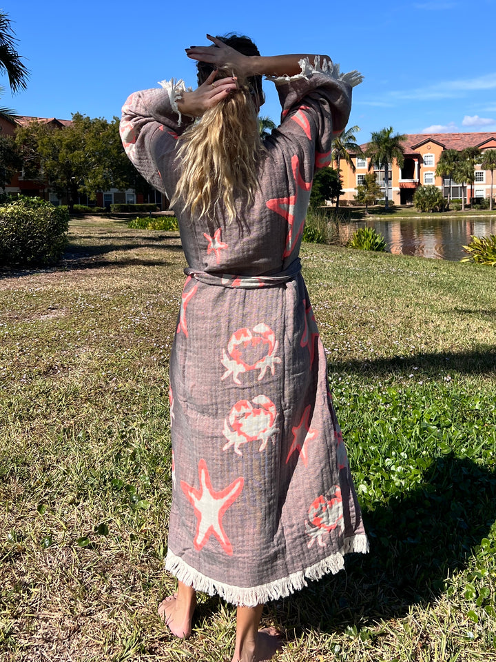 A barefoot person by a pond stands on grass, arms raised, wearing the turkanhome.com Pink Sea Star Robe—a cotton muslin kimono with pink starfish and seashells. Palm trees and colorful houses sit under a blue sky.