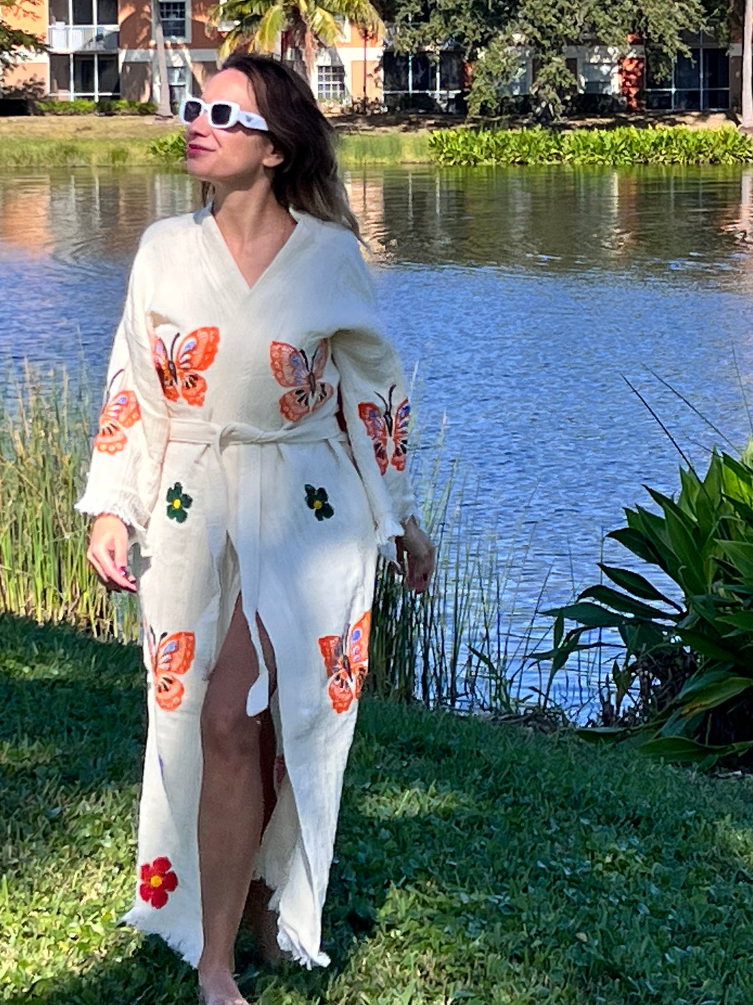 A woman in a Butterfly Kimono Robe-Natural Color by turkanhome.com and sunglasses stands gracefully by the water.