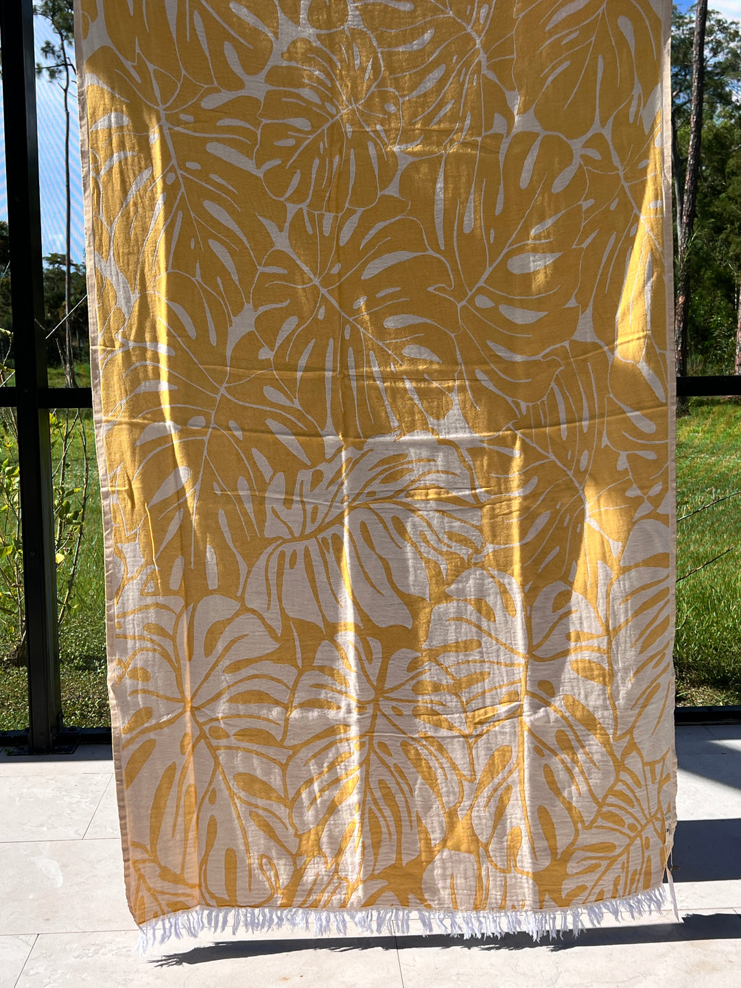 The Botanical Turkish Towel from turkanhome.com, made of 100% cotton with a yellow and white monstera leaf pattern and charming white fringe, hangs in sunlight, with lush greenery visible through a screened enclosure.