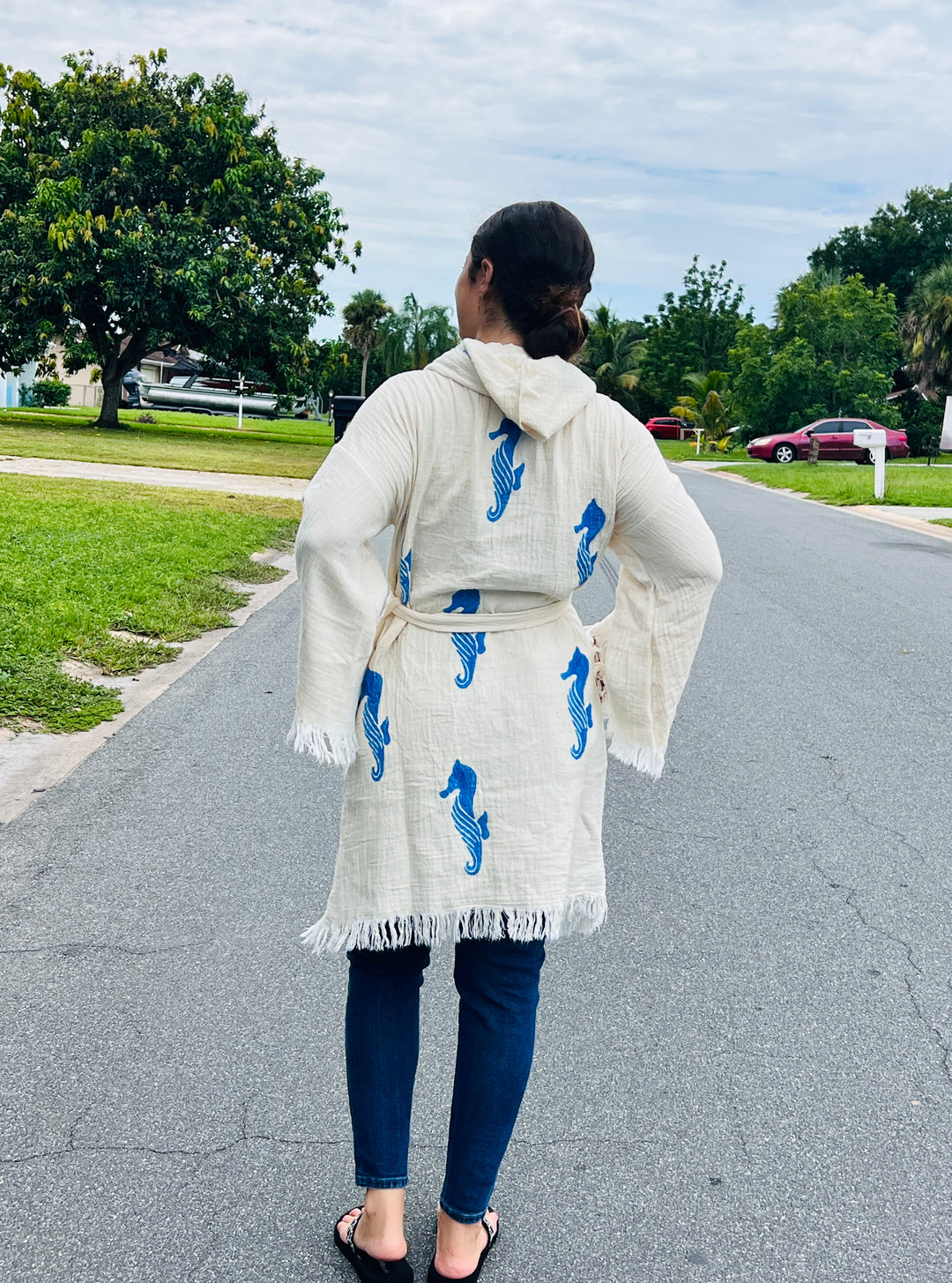 A woman stands on a suburban street wearing the turkanhome.com Seahorse Kimono Robe with Hoodie, paired with jeans and flip-flops. She faces away, with trees, grass, and parked cars in the background.
