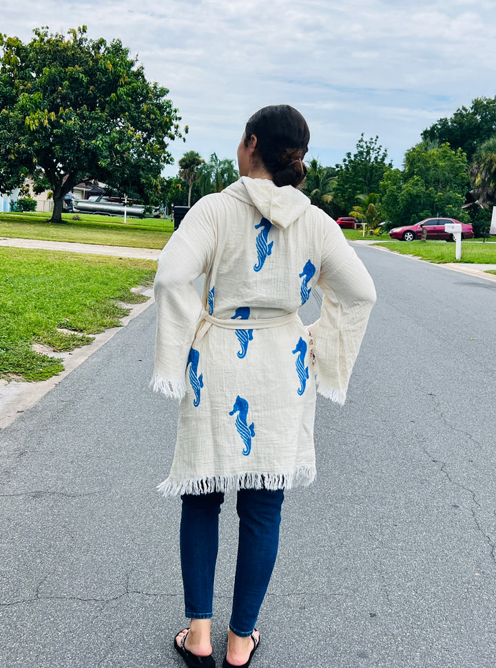 A woman stands on a suburban street wearing the turkanhome.com Seahorse Kimono Robe with Hoodie, paired with jeans and flip-flops. She faces away, with trees, grass, and parked cars in the background.