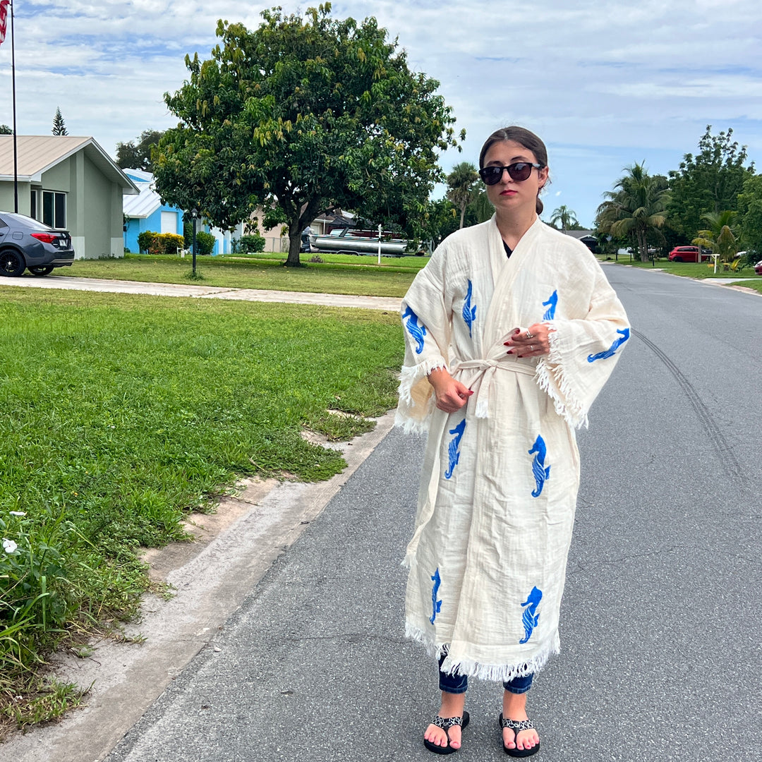 A woman stands on a suburban street in jeans, sandals, and sunglasses, draped in the Seahorse Kimono Robe by turkanhome.com—its blue designs perfect for sunny days. Lush green lawns and trees complete the scene.