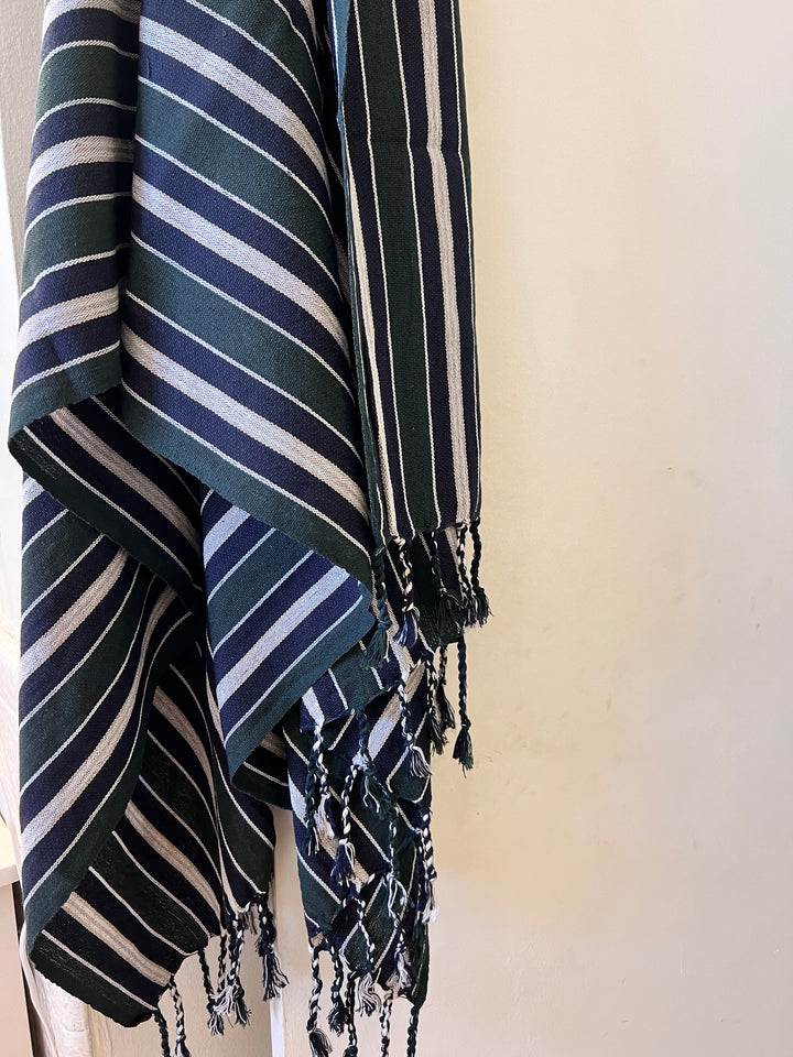 The Halep Handwoven Turkish Towel by turkanhome.com features dark green all-natural fibers, white horizontal stripes, and fringed edges, shown hanging against a light-colored wall. Can also be used as a throw or shawl.