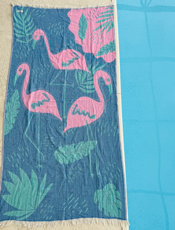 The Flamingo Vibe Towel from turkanhome.com is a reversible, quick drying towel featuring three pink flamingos with tropical leaves on blue, shown on beige tiles by a bright blue pool.