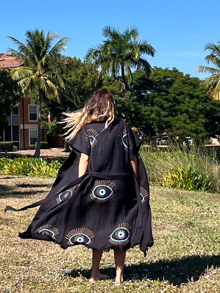 A person with long hair walks barefoot on grass, wearing the Lucky Eye Kimono Robe-Black from turkanhome.com, decorated with bold blue eye motifs. Palm trees and a building appear under the sunny sky in the background.