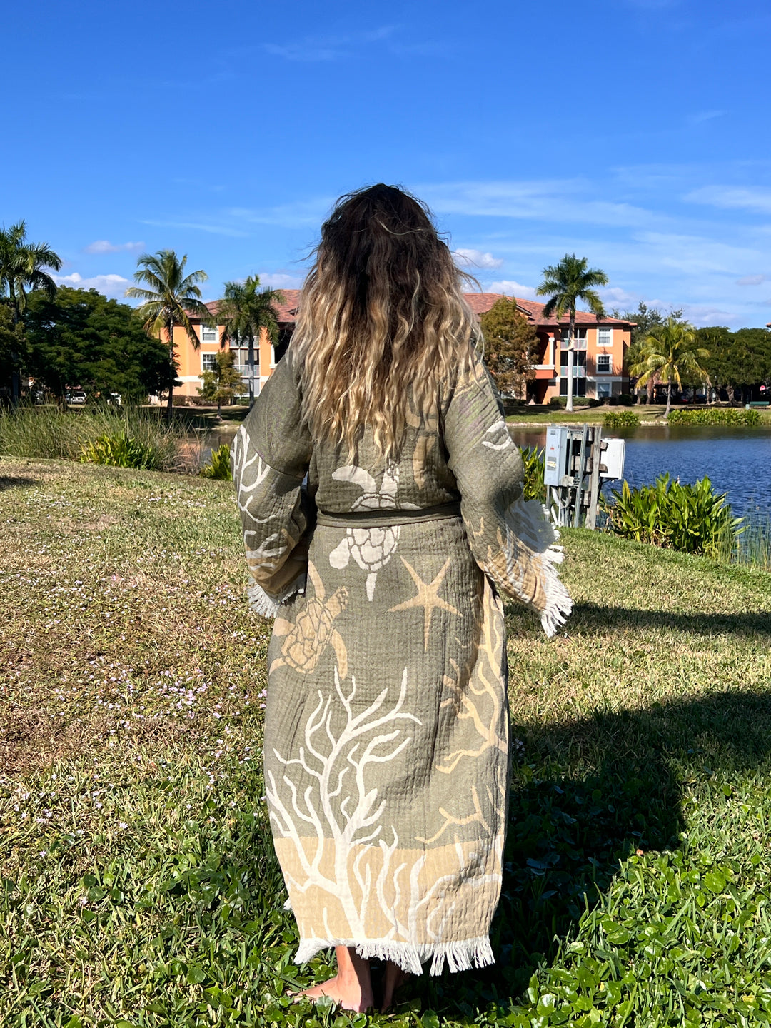 A woman with wavy ombré hair stands barefoot on grass, modeling the Green Turtle Robe kimono beach wear by turkanhome.com. She faces away from the camera, framed by palm trees, a lake, and buildings beneath a blue sky.
