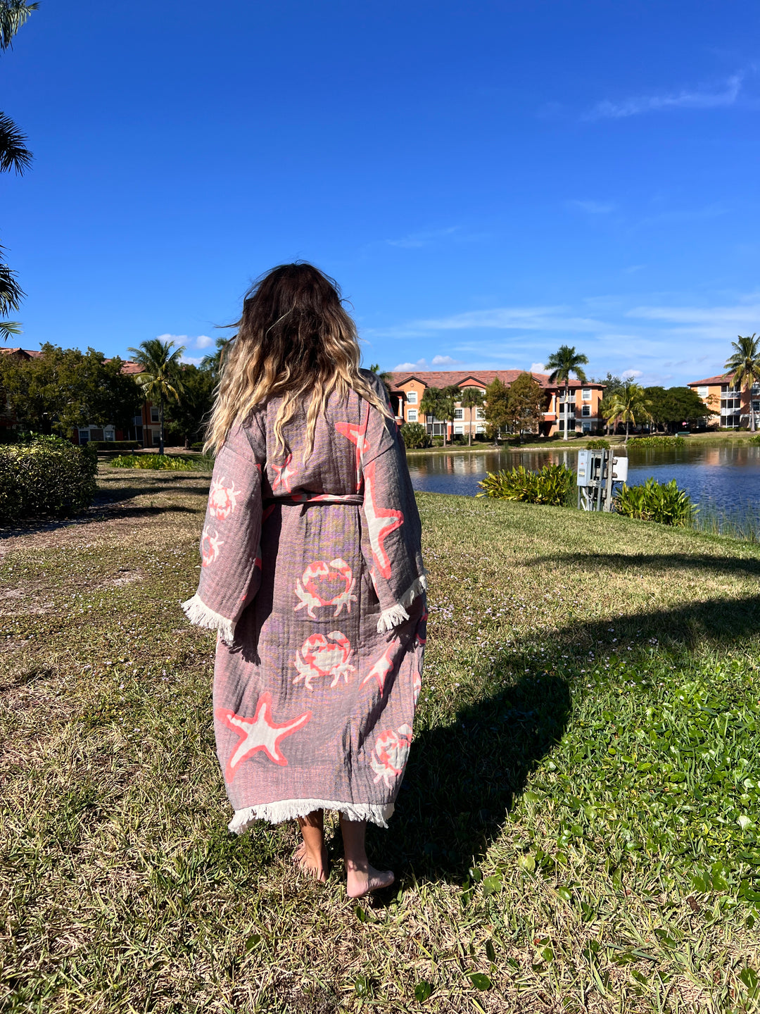 A woman with wavy hair stands barefoot on grass, facing a pond and colorful buildings, wearing the Pink Sea Star Robe Kimono (Beach Wear) by turkanhome.com under a clear blue sky with palm trees around.
