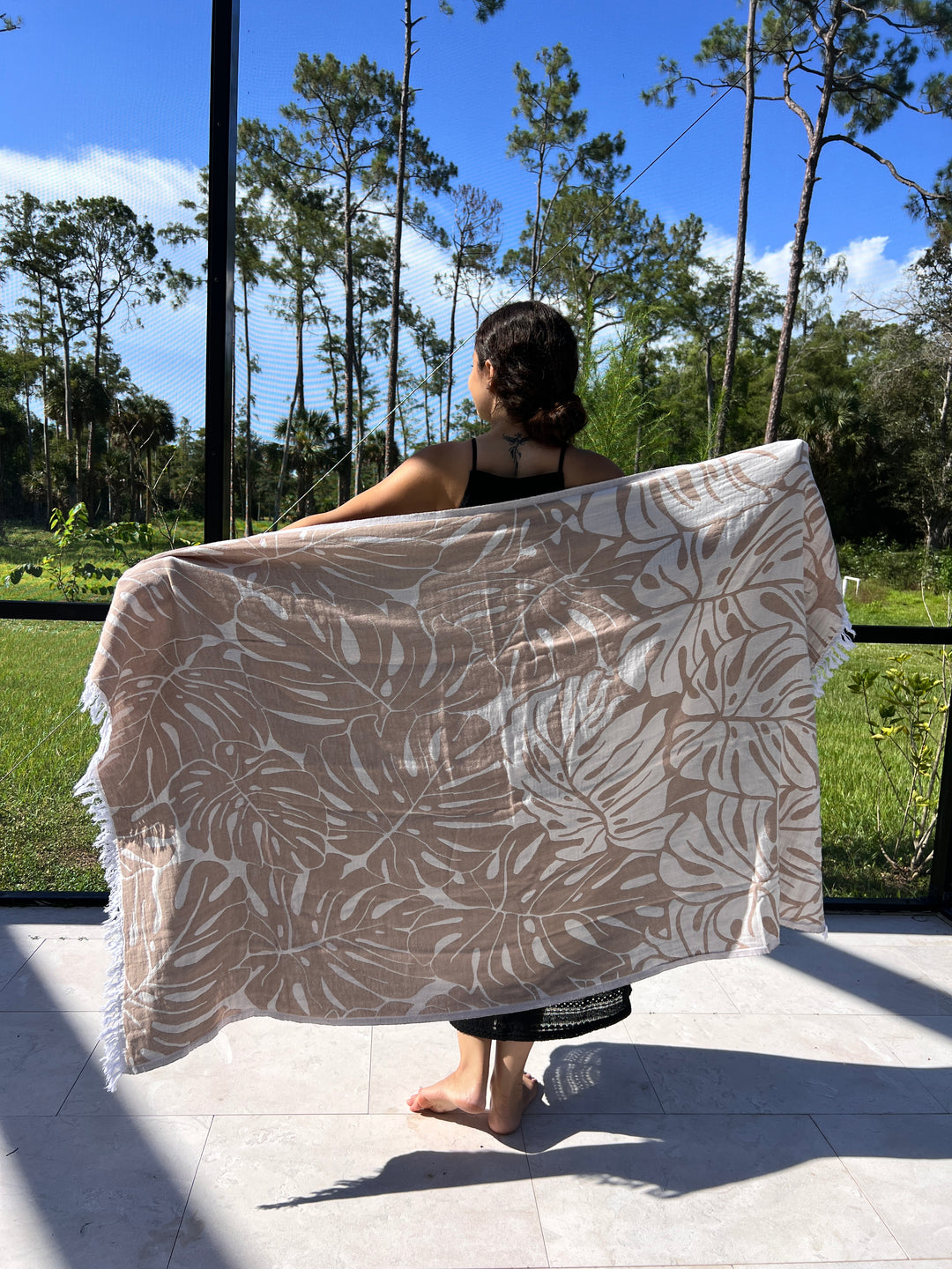 A person stands barefoot on a tiled floor, holding the Botanical Turkish Towel from turkanhome.com with beige and white leaf patterns behind their back, while green trees and grass are visible through large windows in the background.