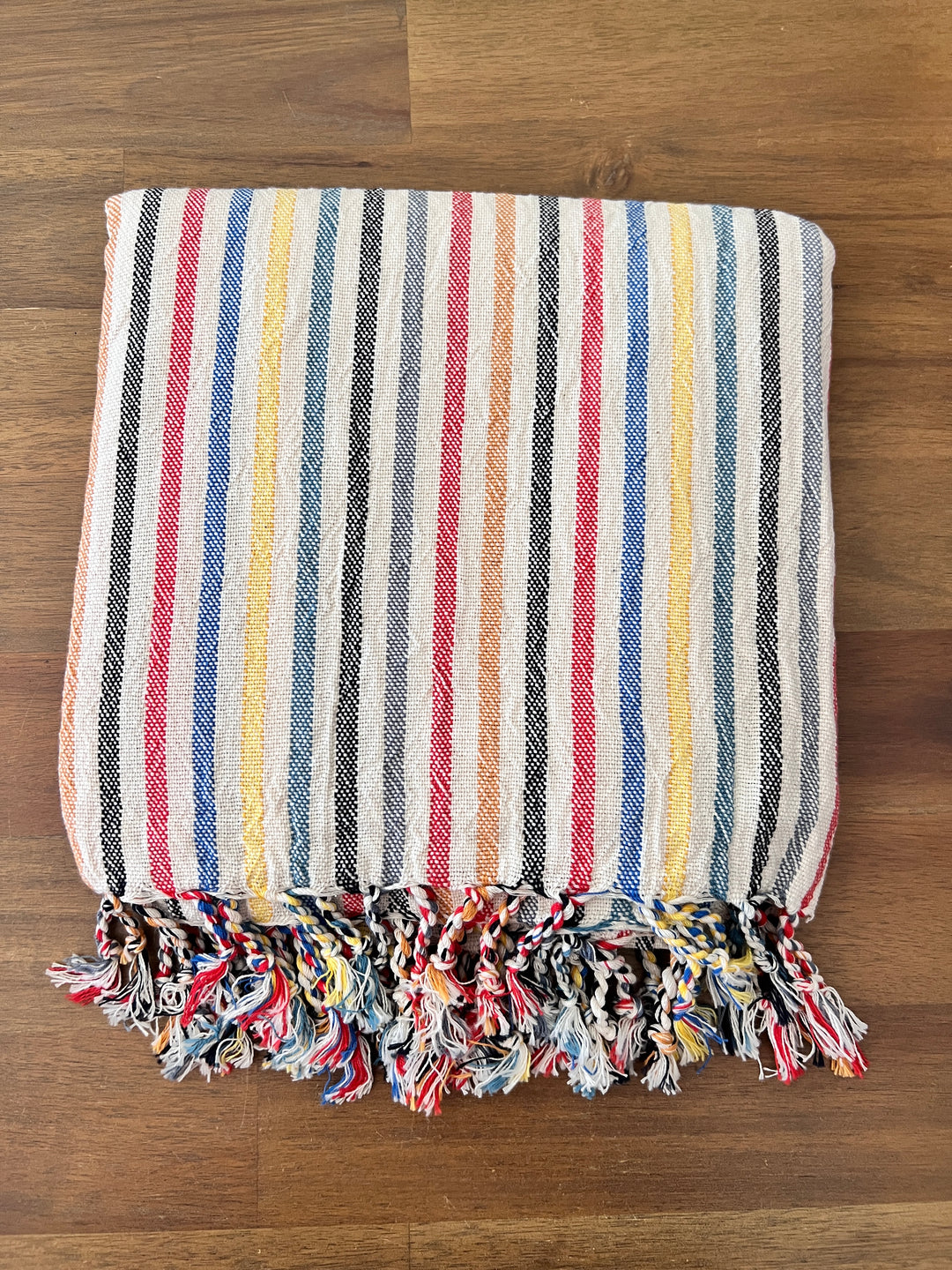 The Mardin Handwoven Turkish Towel, Throw, Shawl by turkanhome.com features colorful blue, yellow, red, black, and white vertical stripes with matching fringes. This folded piece is handcrafted from all-natural fibers and displayed on a wooden surface.