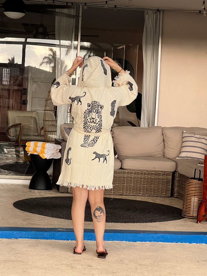 By the pool, a person adjusts the hood of a turkanhome.com Black Leopard Hooded Cotton Kimono Robe. A sofa and patio furniture are in the background, and a large tattoo is visible on their left calf.