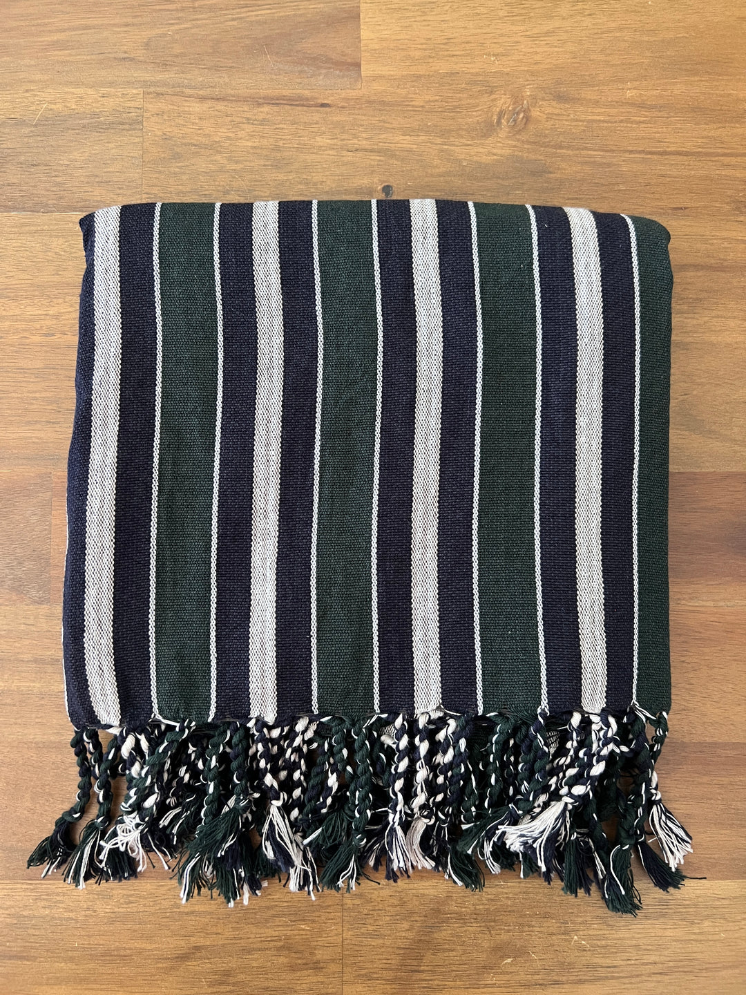 The Halep Handwoven Turkish Towel, Throw, and Shawl by turkanhome.com is handcrafted from all-natural fibers, features dark green, black, and white stripes with twisted fringe tassels, and is displayed folded on a wooden surface.
