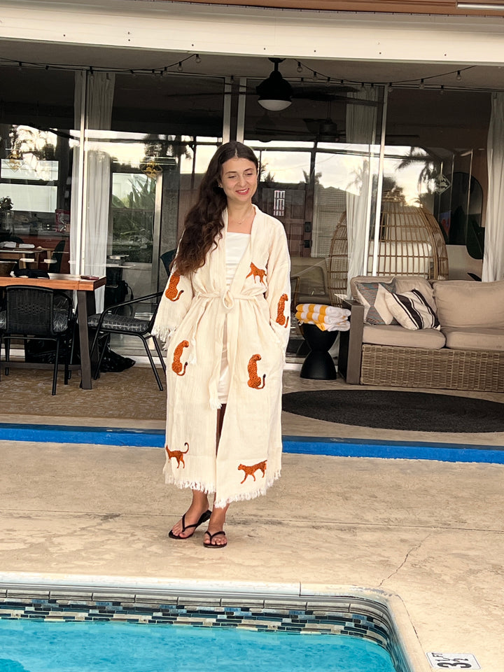 A woman wears the Orange Leopard Cotton Kimono Robe from turkanhome.com as a lightweight boho beach cover up by a pool, paired with sandals. Outdoor furniture and large windows appear in the background.
