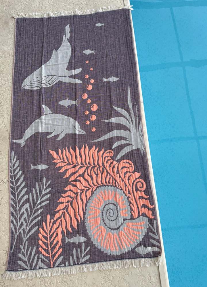The Dolphin and Whale Turkish Towel by turkanhome.com, featuring gray dolphins, fish, coral, and an orange spiral seashell design, is displayed by the edge of a blue swimming pool.