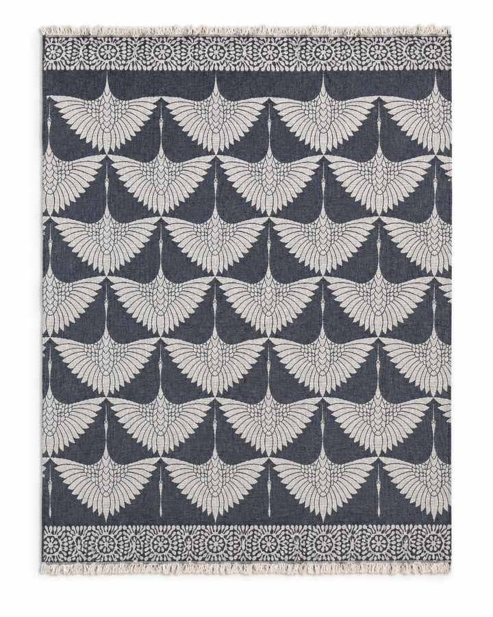 The Crane Muslin Throw Blanket Natural Border–Reversible Turkish Cotton from turkanhome.com features repeating white cranes with outstretched wings set against a dark background. Its rectangular design is complemented by a border adorned with circular and floral motifs, while short fringe on the top and bottom edges adds an elegant finishing touch reminiscent of a classic muslin throw.