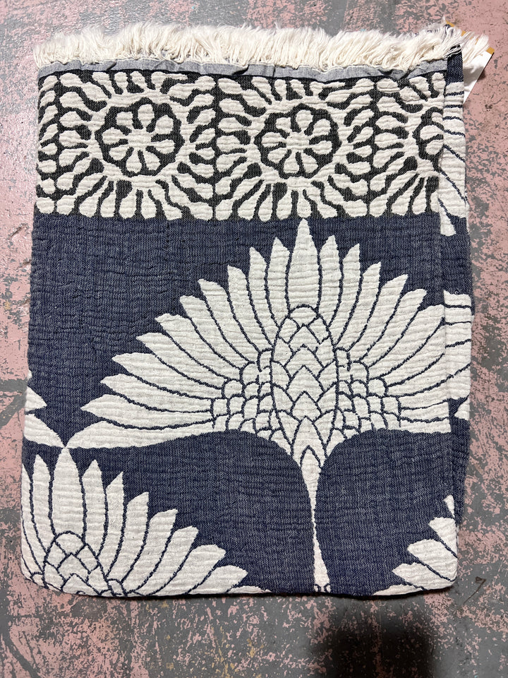 The Crane Muslin Throw Blanket by turkanhome.com is a reversible Turkish cotton throw with a navy and white floral design, showcasing a large central flower, smaller flowers on top, and a fringed border.