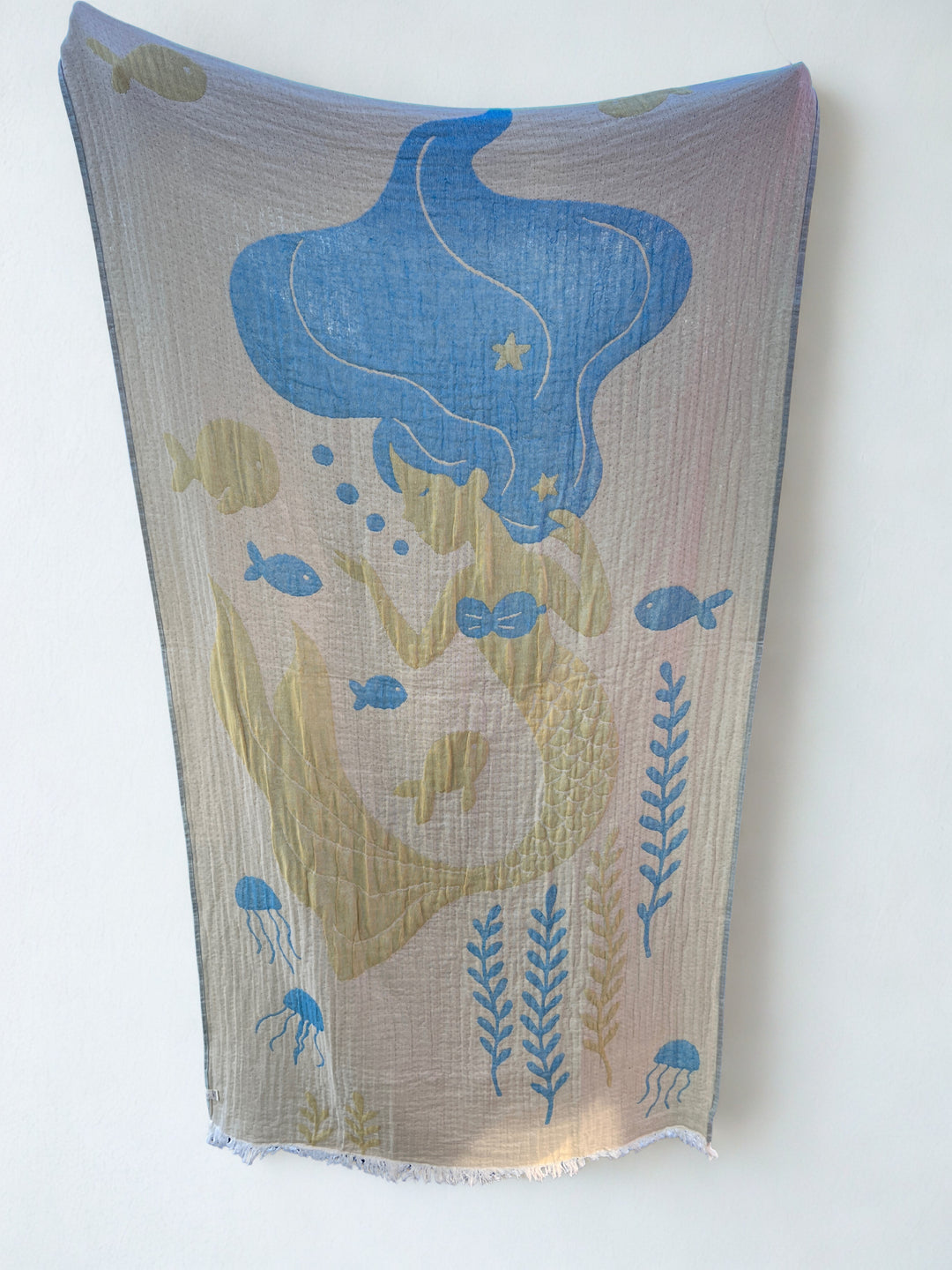 The Mermaid Turkish Towel from turkanhome.com hangs on the wall, showing a blue-haired mermaid with yellow fish, seaweed, and jellyfish in blue and yellow. This quick-drying beach towel features a light background.