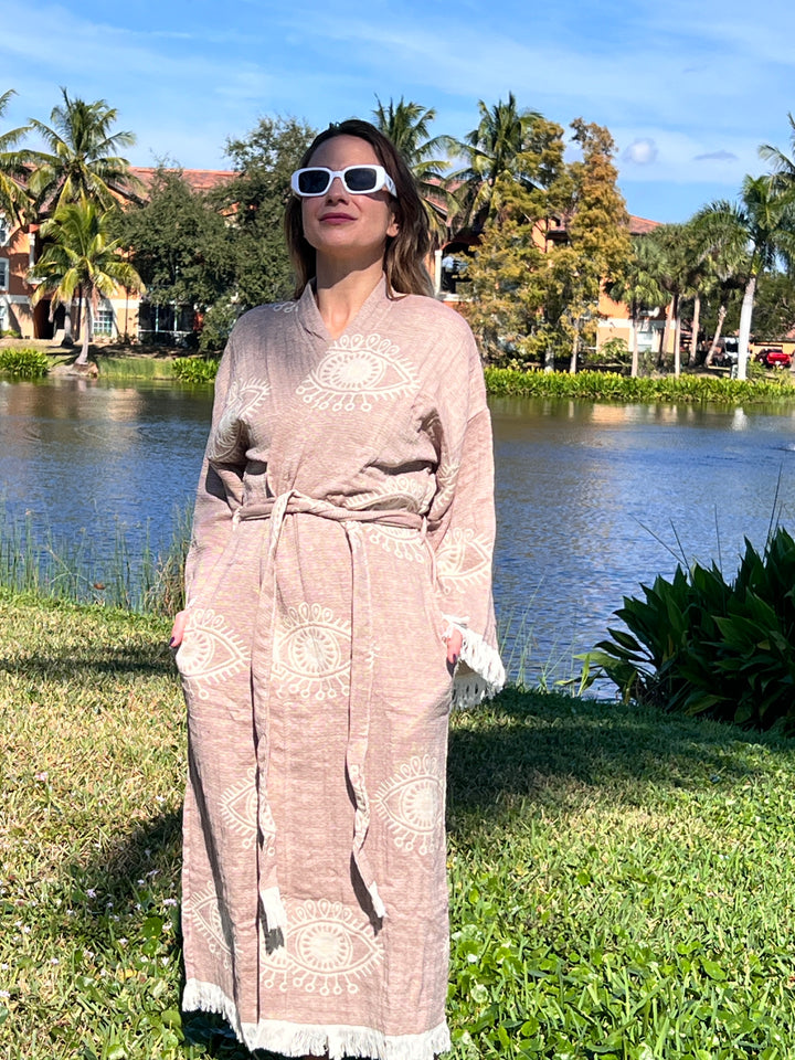 A woman in the Brown Eye Robe Kimono by turkanhome.com stands on grass near a pond, with palm trees and buildings in the background under a blue sky.