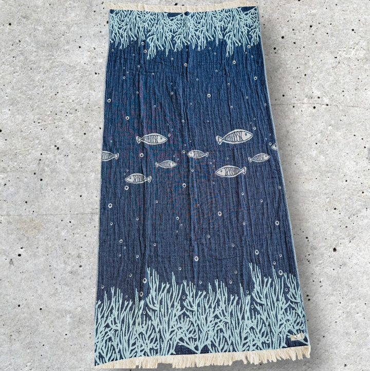 The Fishes and Coral Turkish Towel by turkanhome.com is a blue 100% cotton jacquard weave with white fringe, featuring swimming fish and coral on a dark ocean background, shown laid flat on a light speckled surface.