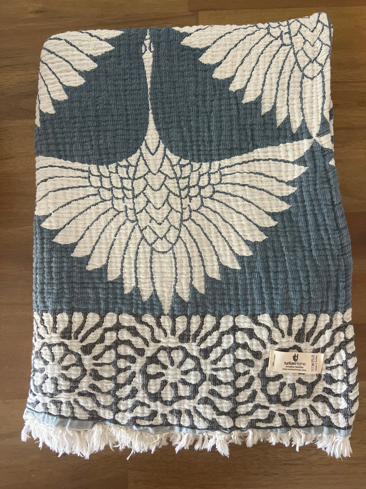The Crane Muslin Throw Blanket – Reversible Turkish Cotton from turkanhome.com, in folded blue and white with a striking crane design and geometric border, is displayed on wood with a fabric tag in one corner.