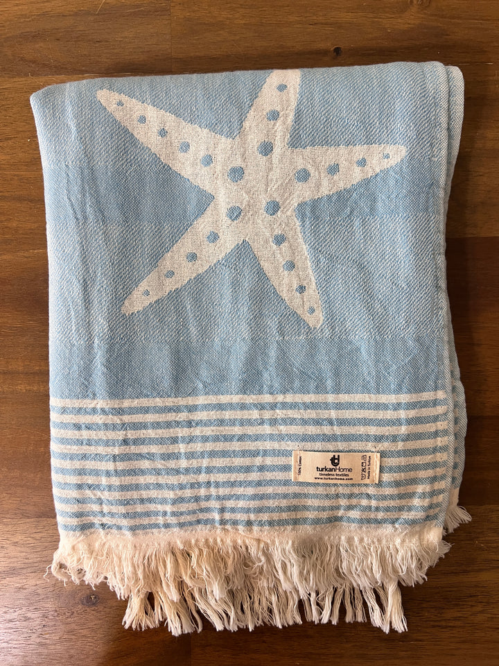 The Sea Star Turkish Towel by turkanhome.com, Jacquard woven from 100% cotton with blue and white starfish and stripe designs plus fringed edges and a fabric label, is folded on a wooden surface.